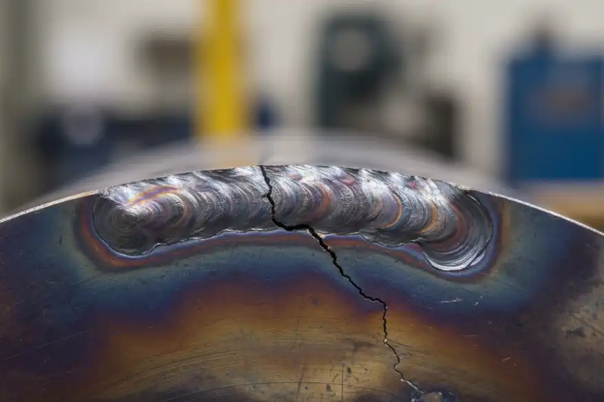 A macro photography shot of a steel weld joint showing a microscopic crack starting specifically in the Heat Affected Zone (HAZ) right next to the weld bead. The metal surface shows thermal discoloration (heat tint). Industrial quality control context, high resolution.
