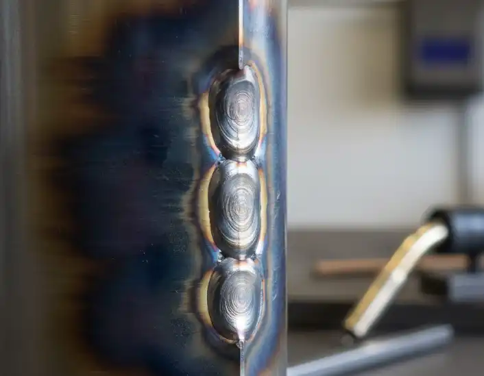 A close-up macro photograph of a stainless steel weld seam. Half of the weld seam is heavily oxidized, showing dark black and dark blue heat tints, while the other half is perfectly clean, shiny, and metallic. Industrial welding context, high resolution, soft lighting.