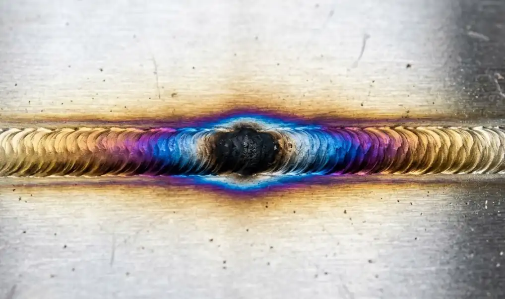 A highly detailed, macro photograph of a stainless steel TIG weld showing a colorful heat tint spectrum. The colors transition smoothly from light pale yellow or straw gold on the outer edge, to deep blue, purple, and finally a harsh burnt black at the very center. Clean, industrial manufacturing aesthetic.
