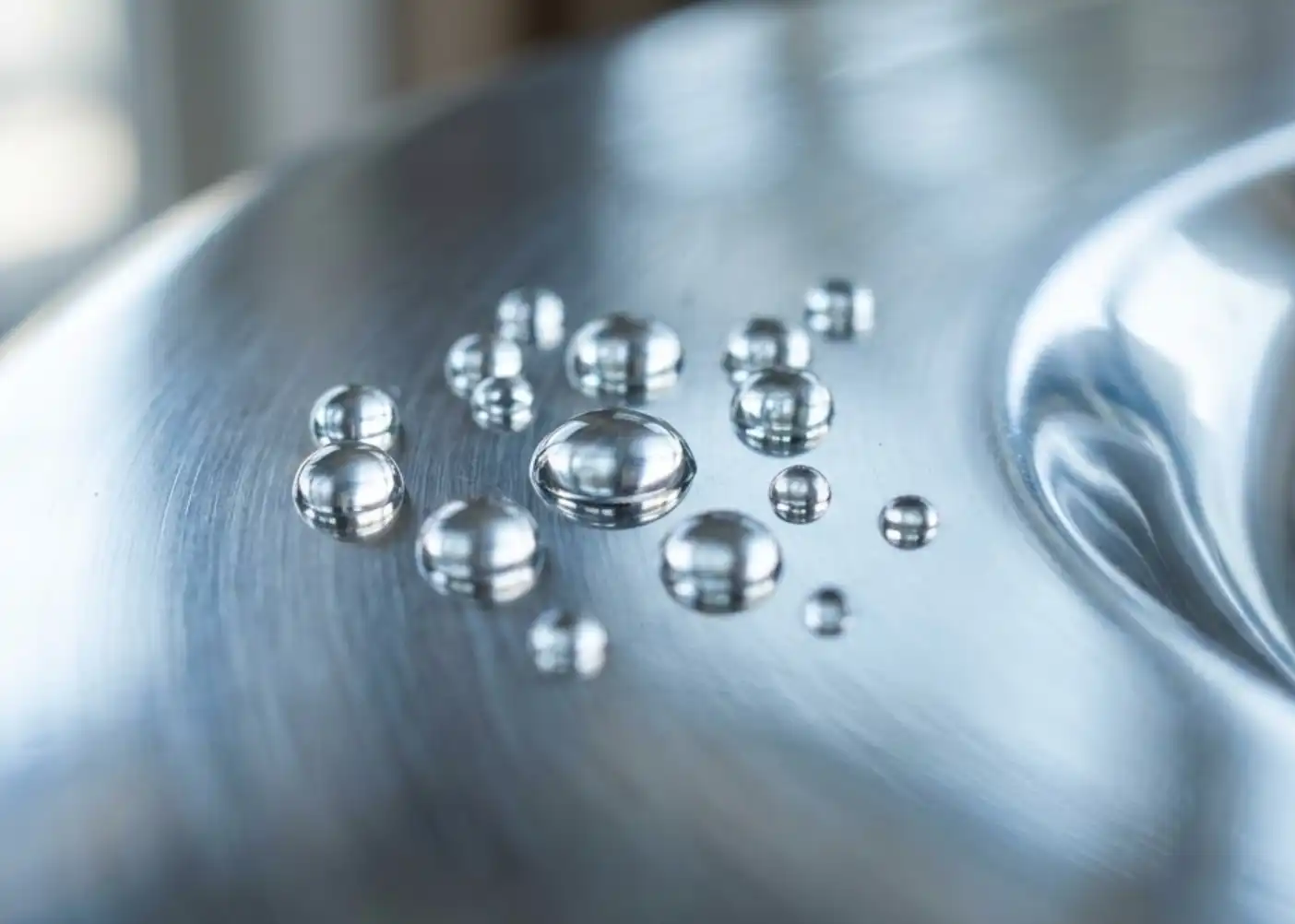 A close-up, high-quality macro photograph of water droplets resting perfectly on a polished, incredibly shiny and rust-free stainless steel surface. The lighting highlights the clean, sleek, and highly reflective nature of the metal.