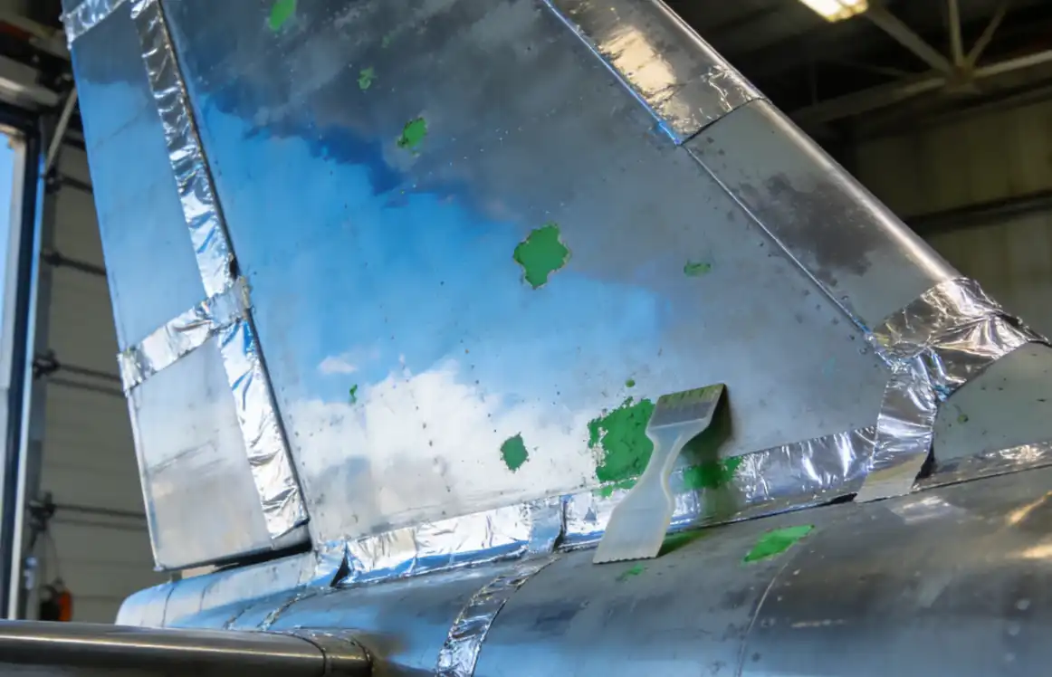 A detailed shot of an airplane stabilizer after a complete stripping process. Most of the surface is clean, reflecting the blue sky from the hangar door, but a few small, stubborn patches of old green paint remain. A plastic scraper lies next to them, indicating the troubleshooting stage. The surrounding bondlines are covered with silver foil tape. High contrast and sharpness.