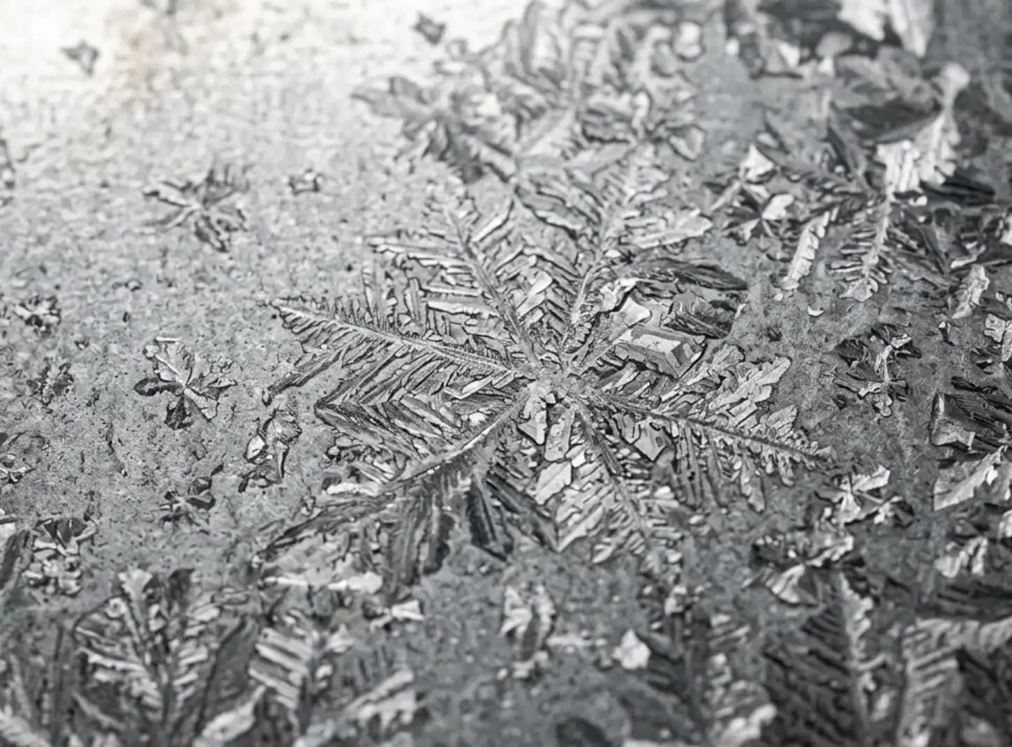 A macro photography close-up of a hot-dip galvanized steel sheet surface. The image clearly shows the distinct, crystalline "spangle" pattern that looks like frost or snowflakes on a matte gray metallic background. High resolution, industrial lighting.