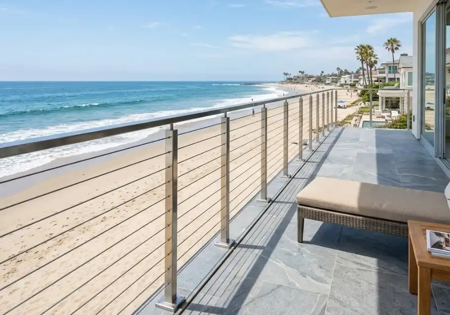 A professional architectural photograph of a sleek, modern stainless steel railing system installed on a beachfront property balcony. The bright, polished metal contrasts with the ocean and sandy beach in the background. High-end coastal construction aesthetic.